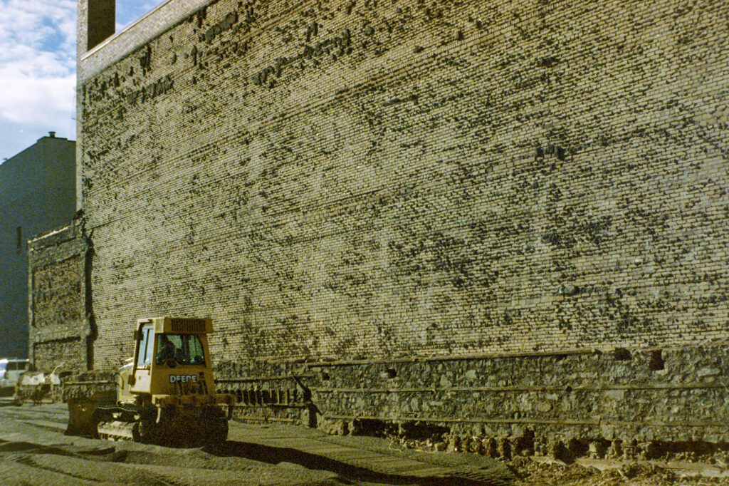 colour photo of sunlit brick exterior wall with bulldozer grading gravel alongside