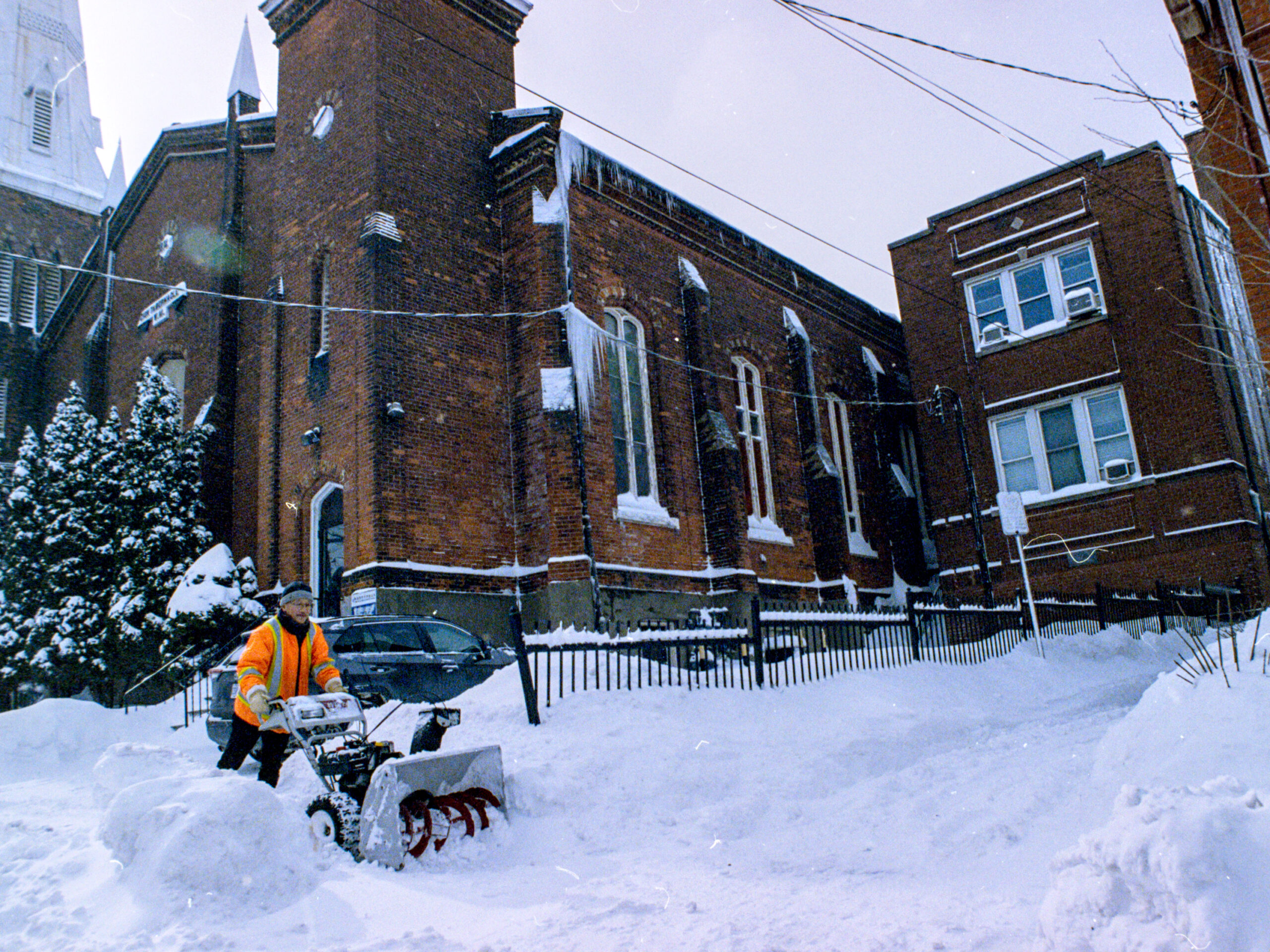 colour photo of man operating snow blower on sidewalk in front of church buildings.