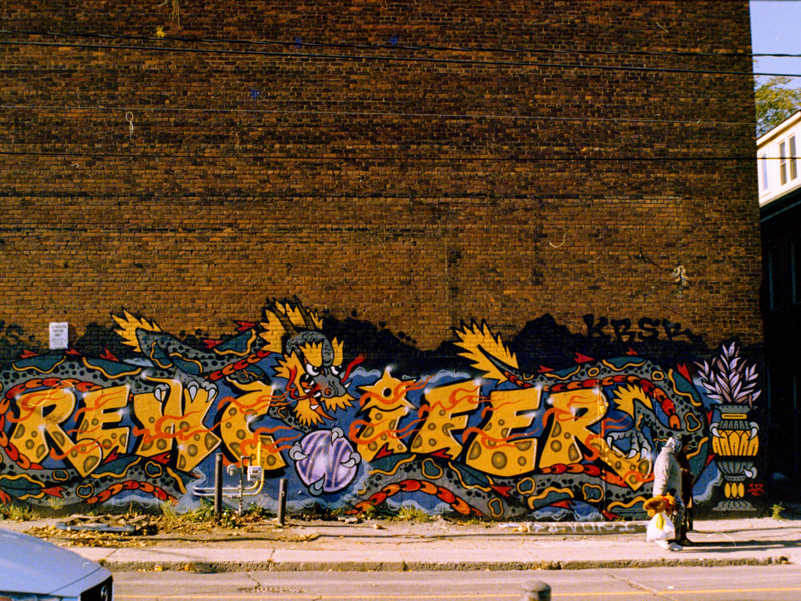 colour photo of woman on sidewalk walking past a windowless brick wall with colourful graffiti