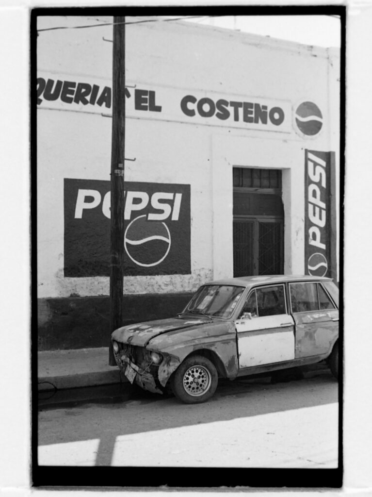 black and white photo of dilapidated car in front of building covered with hand painted pepsi signs