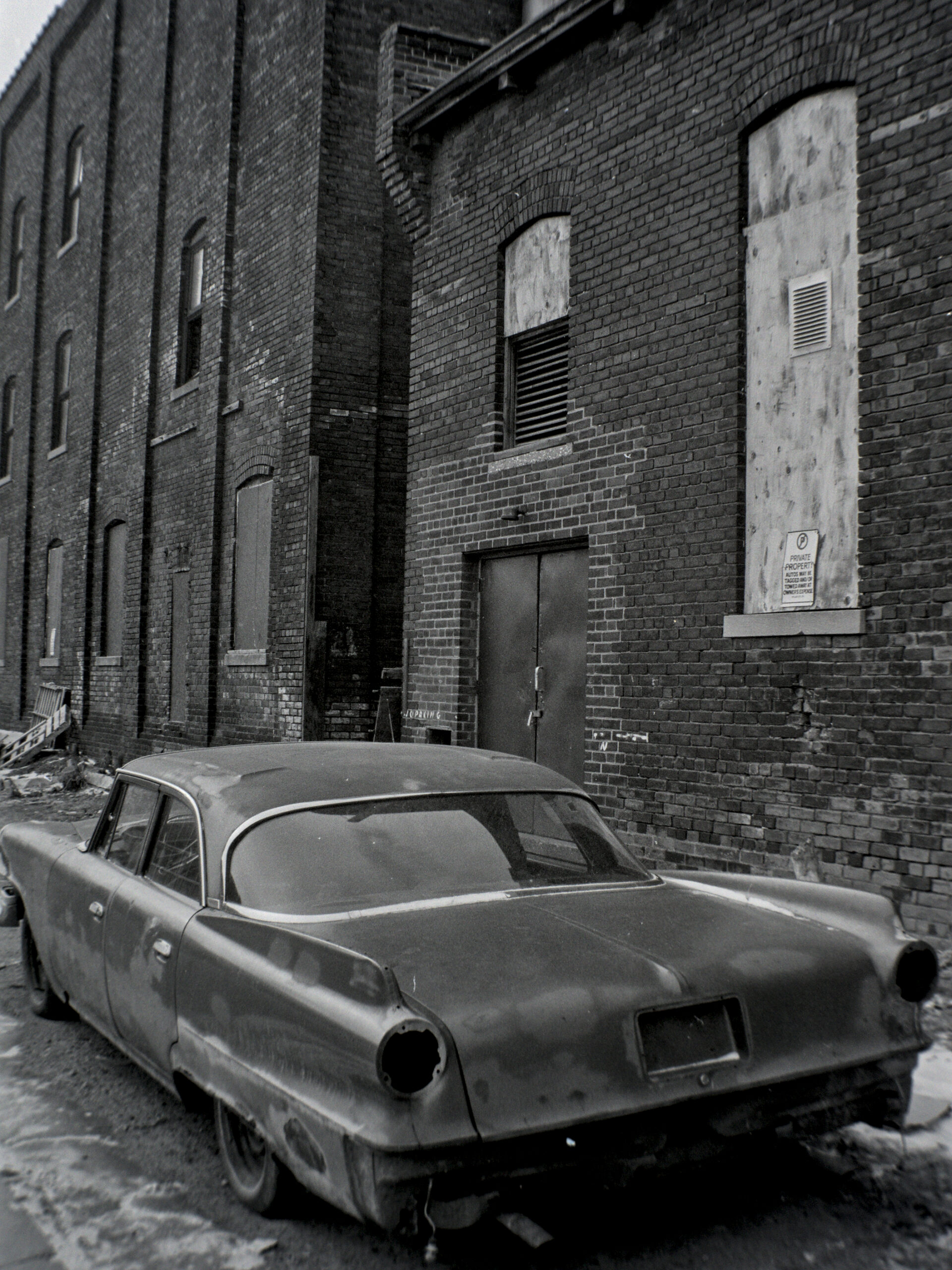 black and white photo of beat up classic sedan parked alongside an old industrial brick building