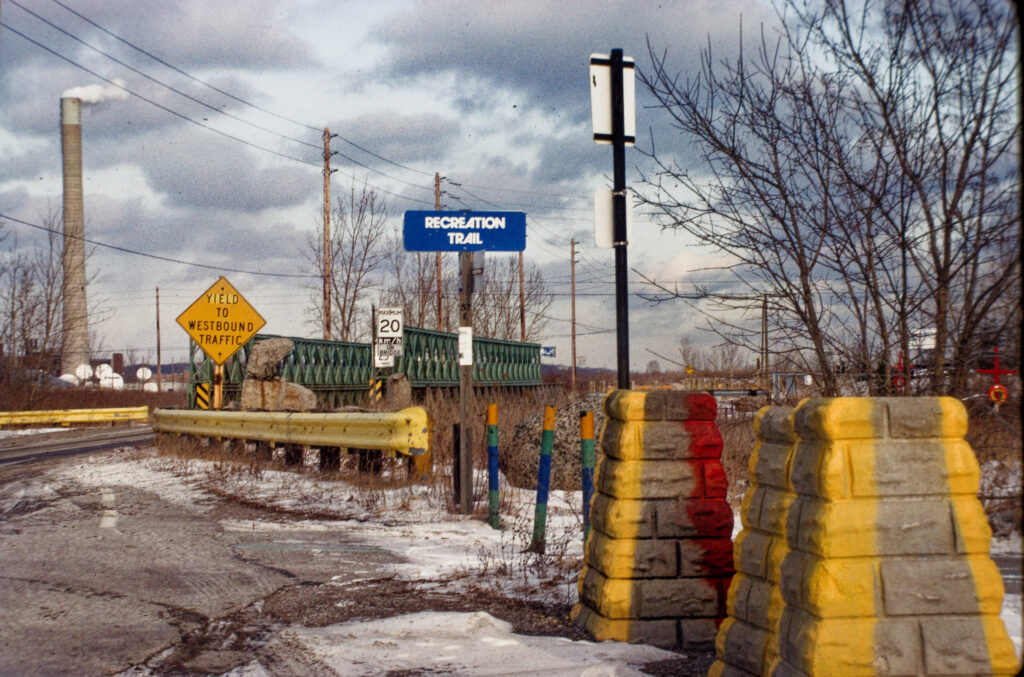 colour photo in winter of various brightly painted bollards and guardrails with bridge, smokestack, trees, and power lines in background
