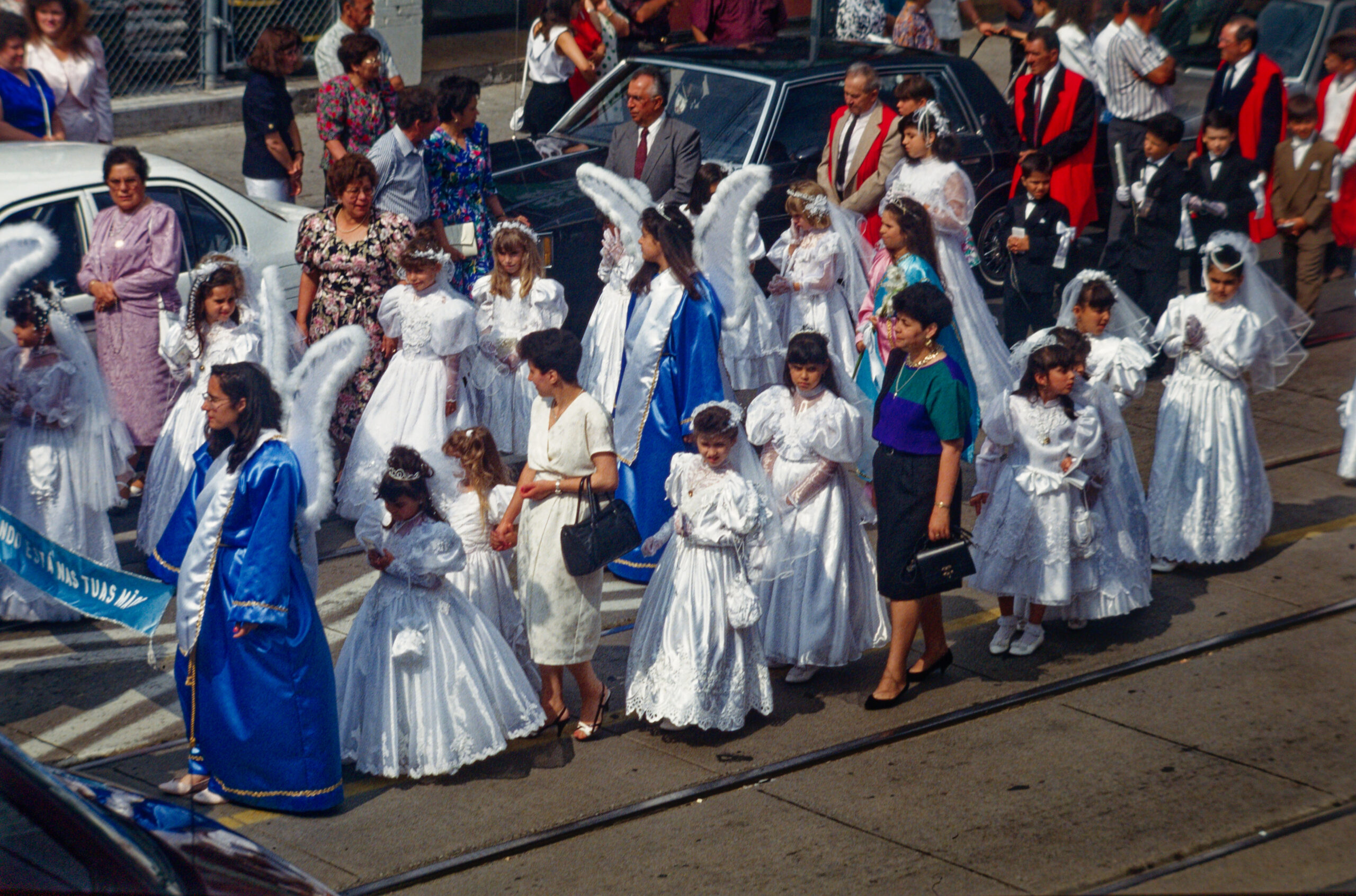 colour photo of girls in fancy white dresses, some with angel wings, moving along the street as part of a parade