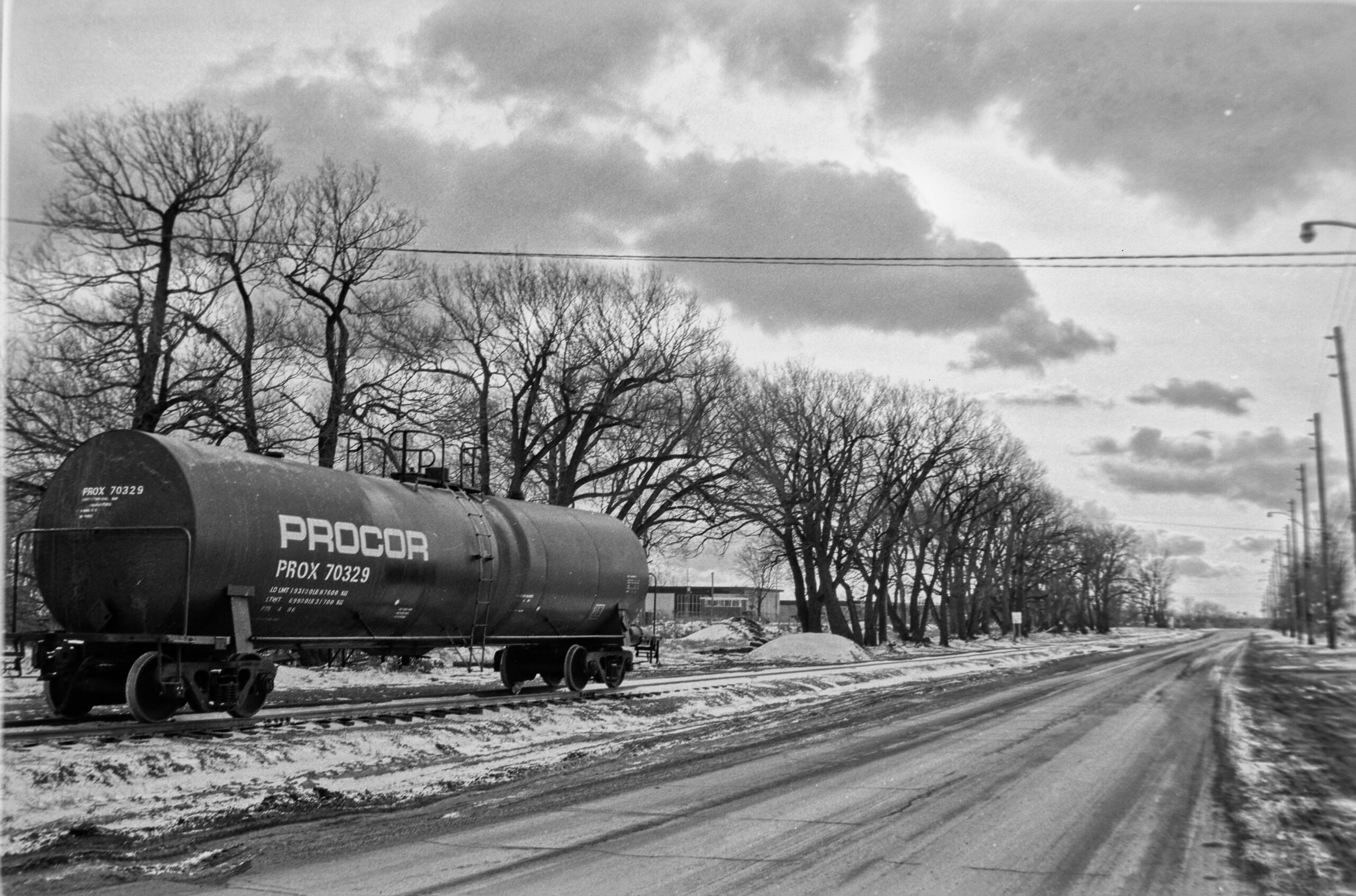 black and white photo of a rail tanker car alongside a tree lined roadway in winter