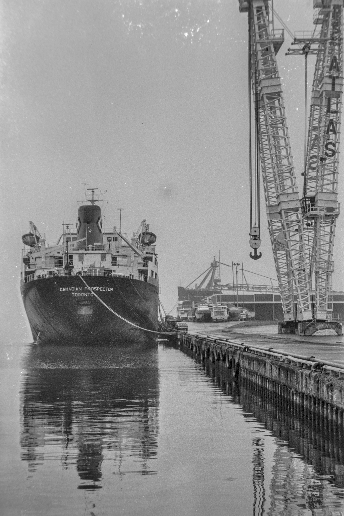 black and white photo of a cargo ship moored alongside a large crane. Ship name on stern "Canadian Prospector, Toronto". Lettering on crane "ATLAS".