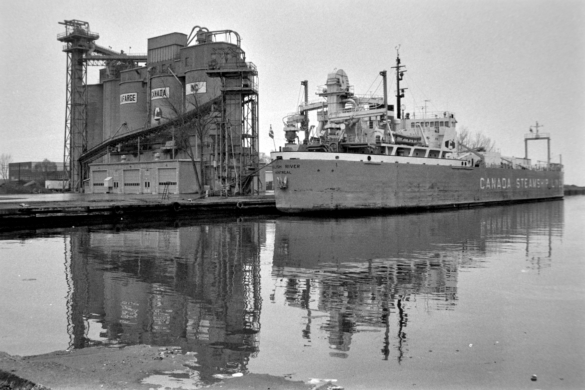 black and white photo of a cargo ship moored alongside cement silos. Ship name on stern "English River, Montreal". Lettering on silos "Lafarge Canada".