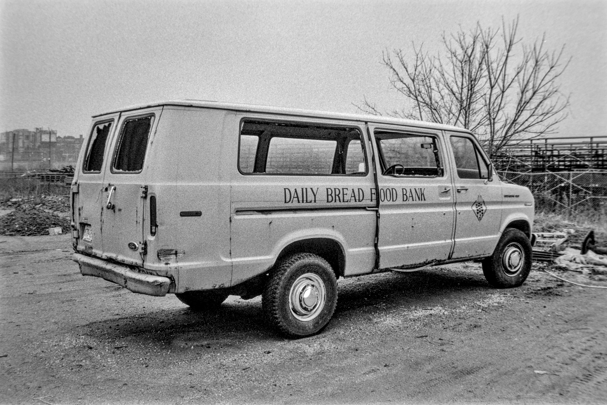 black and white photo of a parked cargo van with windows smashed out. Reads "Daily Bread Food Bank" on the side.