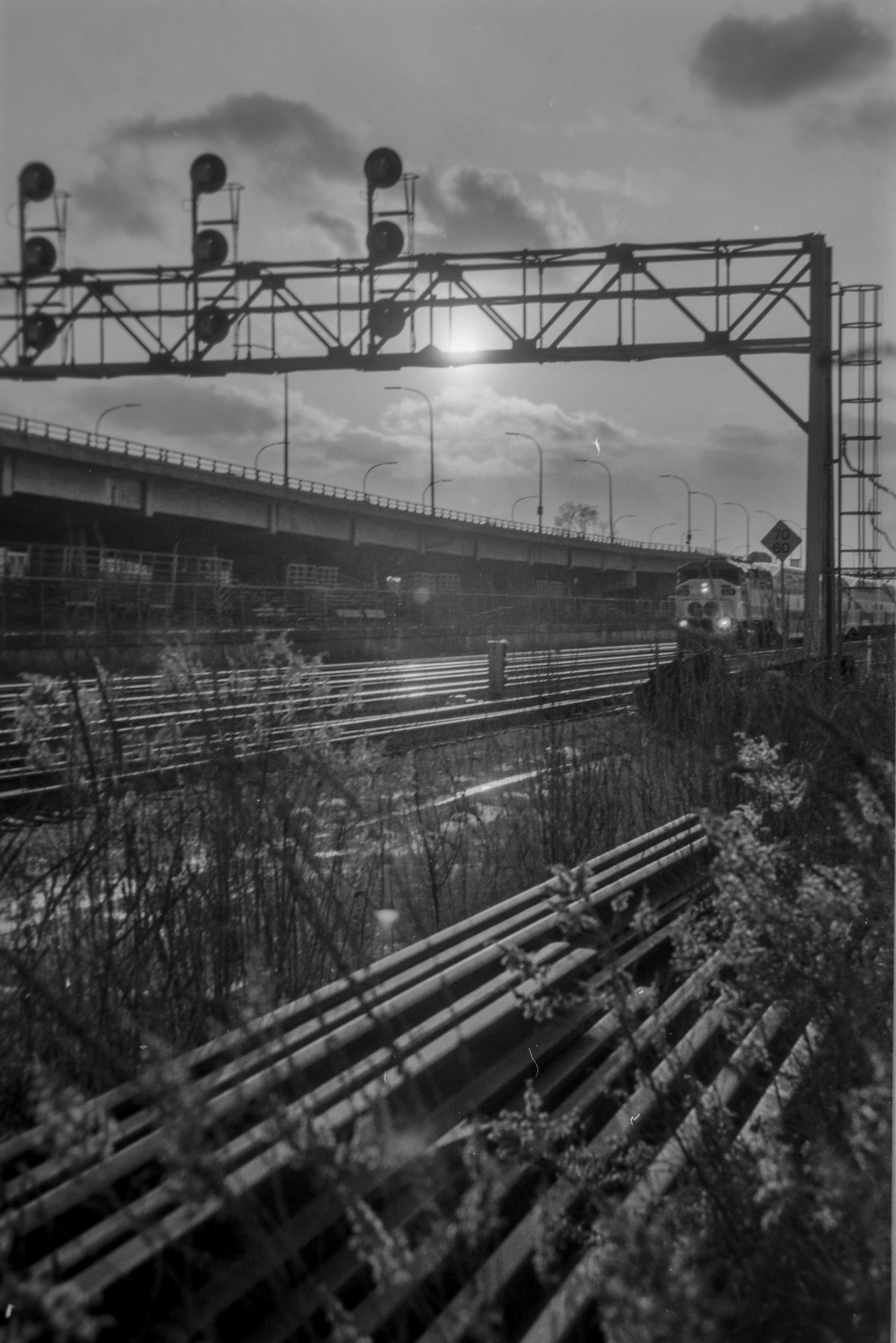 black and white photo of a rail corridor with passenger train approaching. Elevated highway in background.