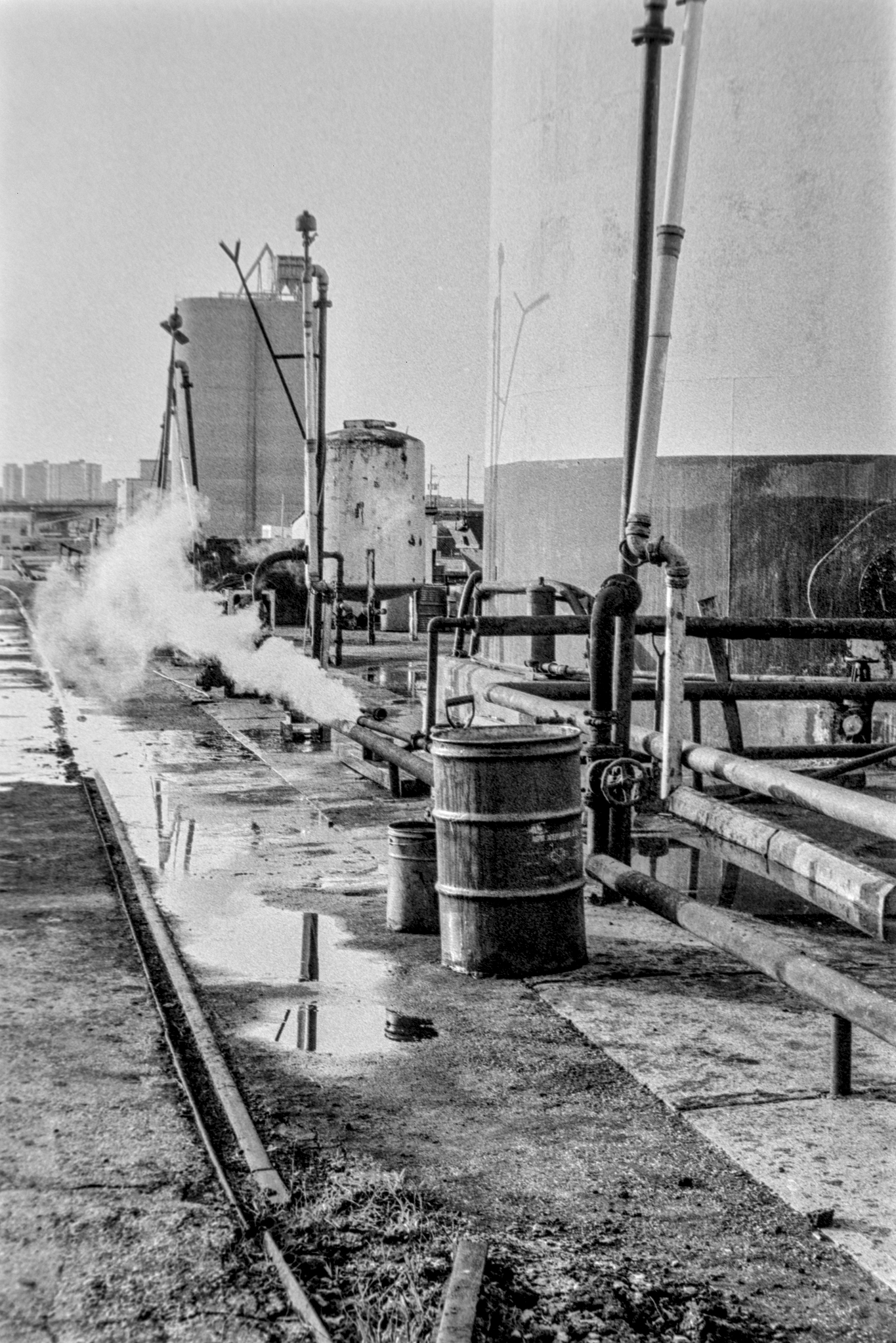 black and white photo of large storage tanks with pipes emitting steam and barrels