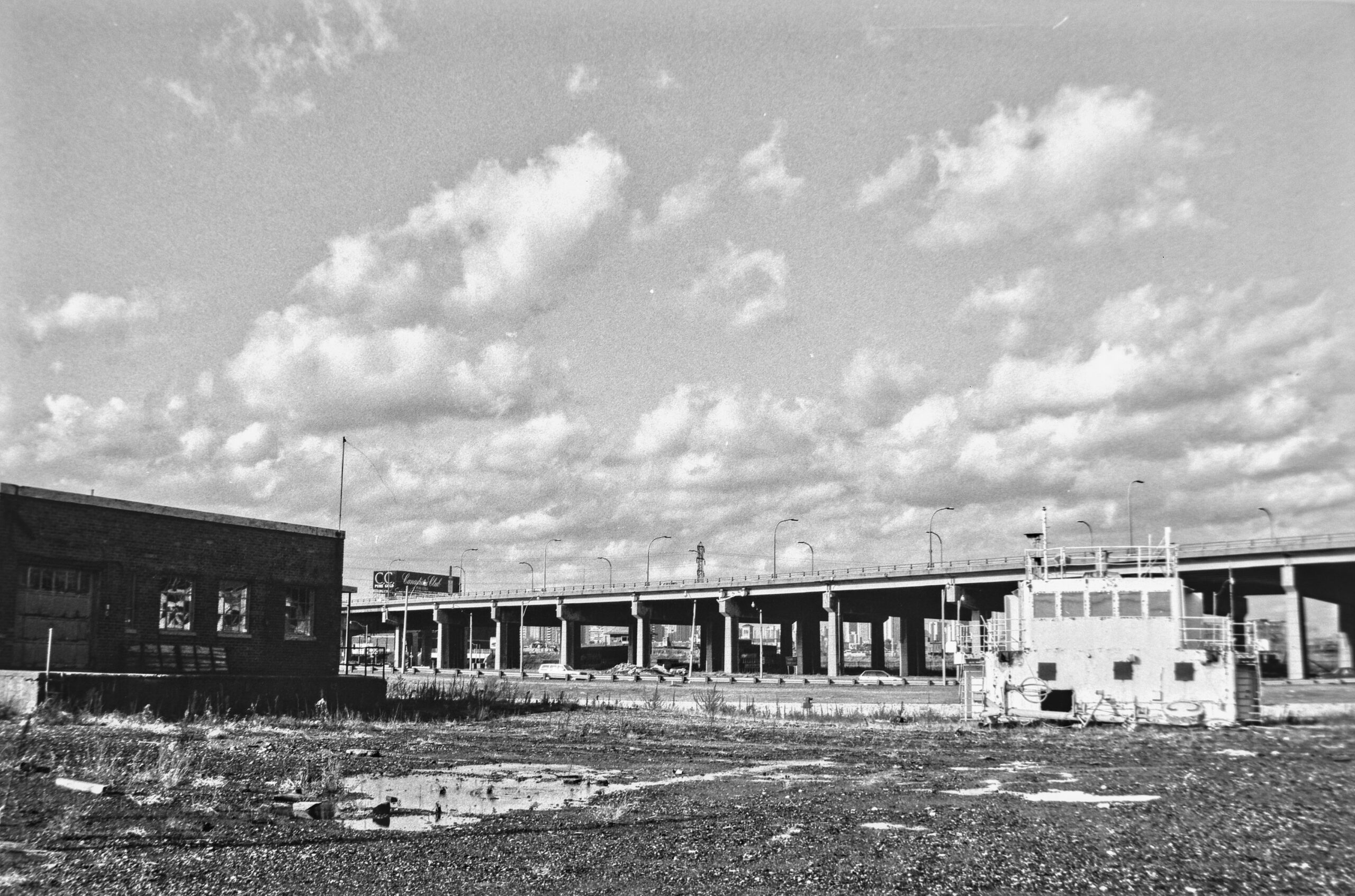 black and white photo of a vacant lot with elevated highway and clouds in background