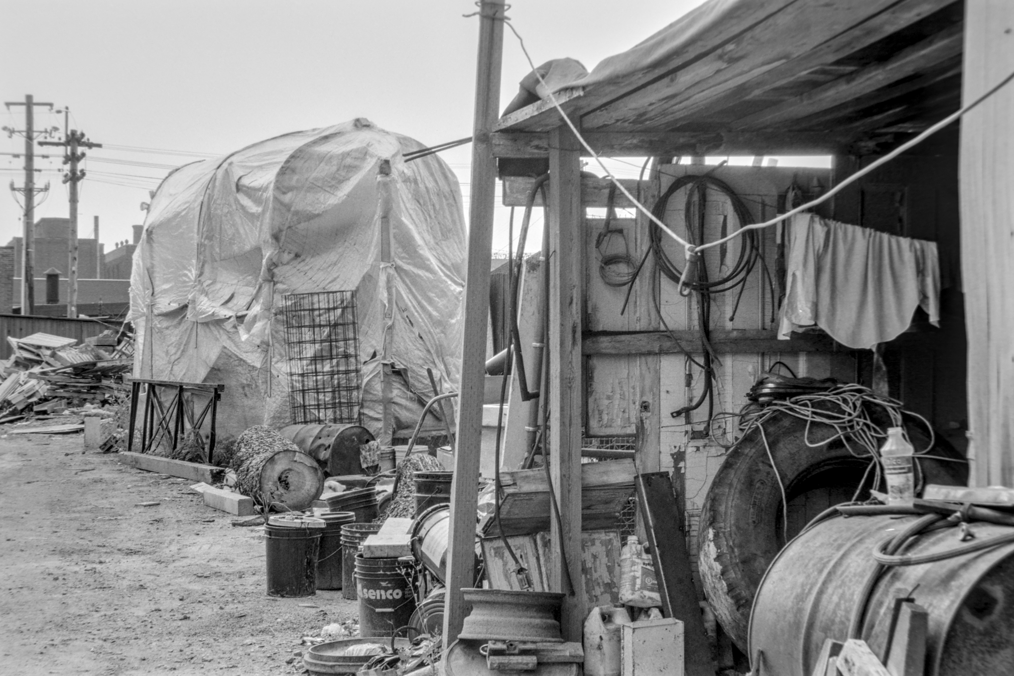 black and white photo of an industrial yard with shed, tools, tires, pails, tarps, and fuel tank.