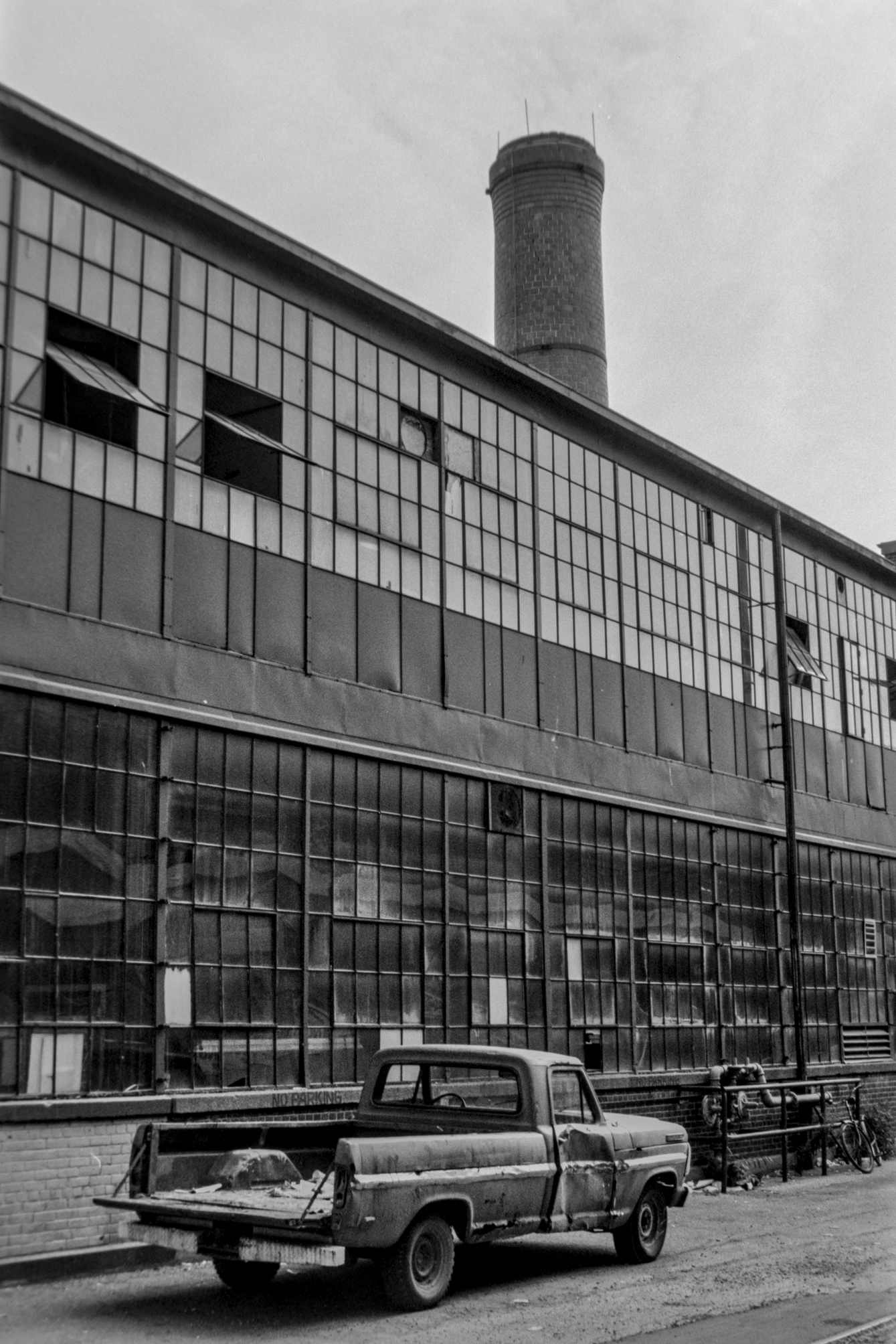 black and white photo of beat up pickup truck parked alongside old industrial building with smokestack behind