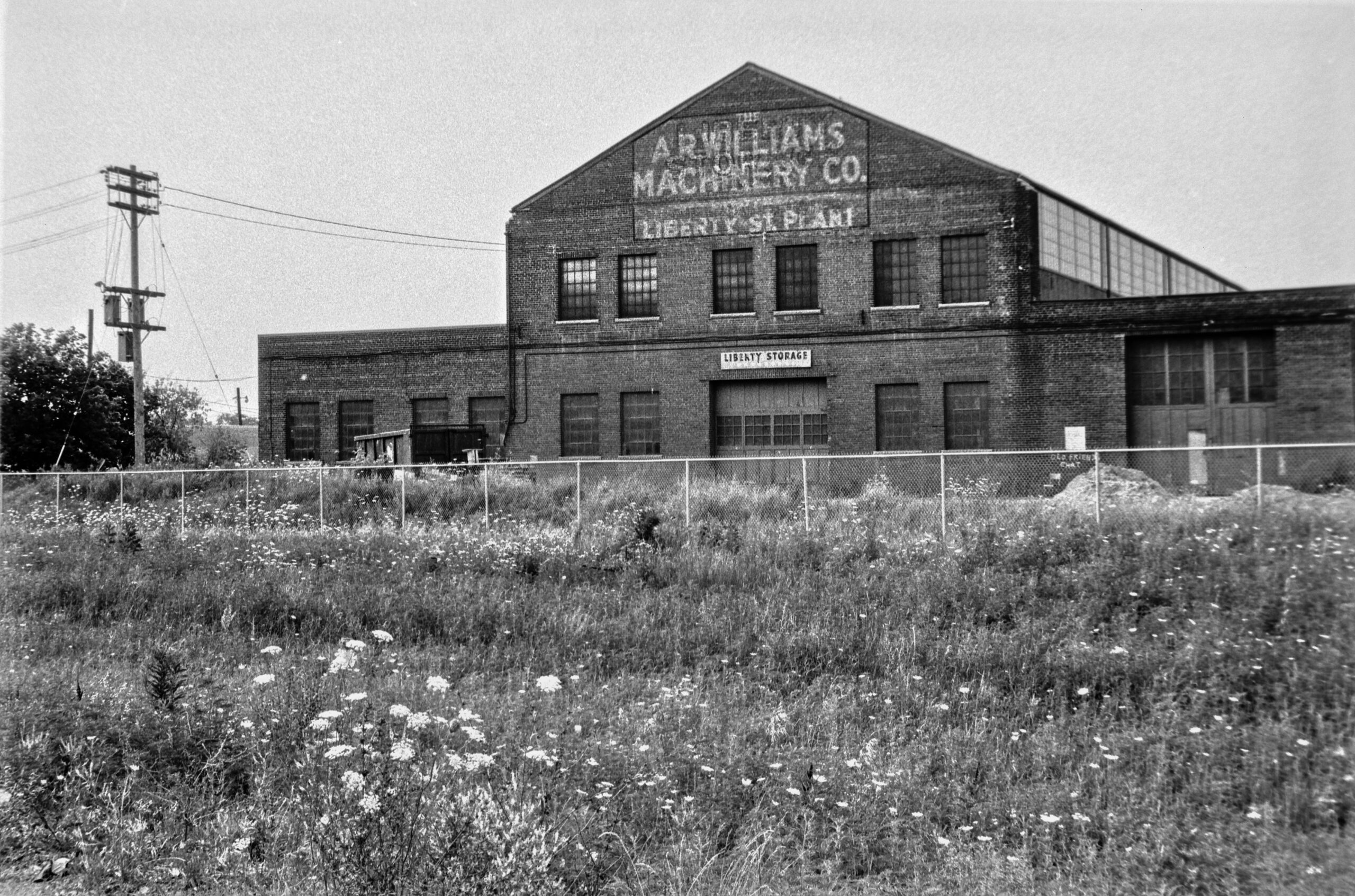 black and white photo of historic industrial building. fading sign reads "A. R. WILLIAMS MACHINERY CO." and "LIBERTY ST. PLANT". Grassy field with wildflowers in foreground.